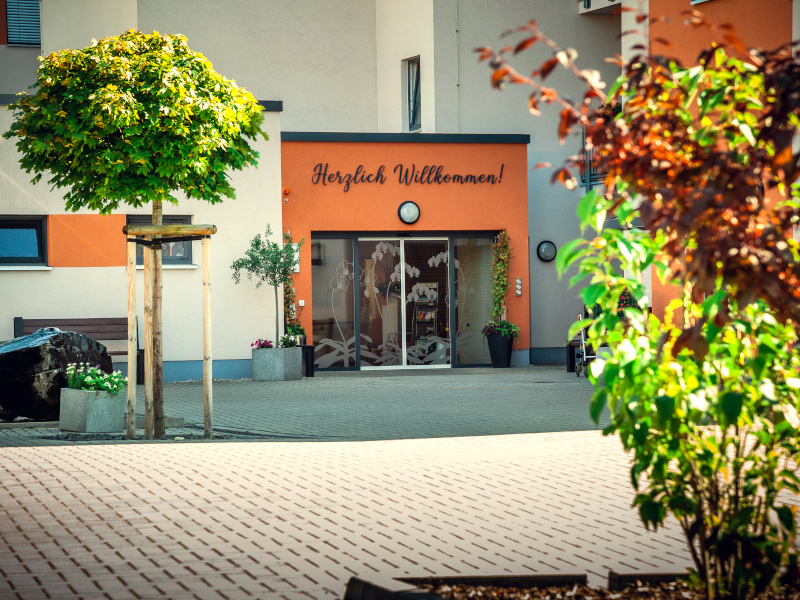 Außenansicht weiß orangenes Gebäude mit Herzlich Willkommen über der Glastür und Außenbegrünung