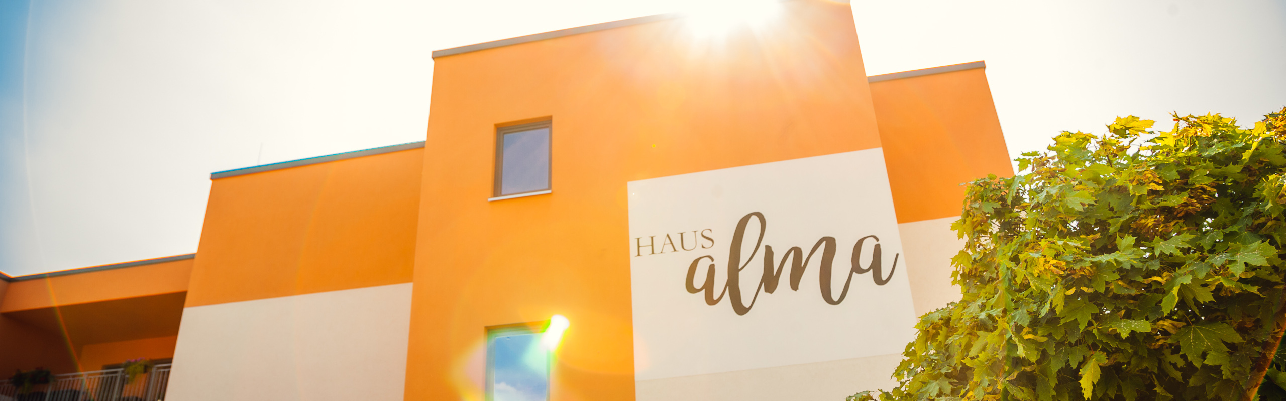 Weiß orangefarbenes Haus im Sonnenschein mit Aufschrift Haus alma und Ahornbaum im Vordergrund